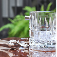The Saint-Louis/Hermes Manhattan Ice Bucket, filled with ice cubes, sits on a reflective surface next to metal tongs, with softly blurred green ferns in the background.