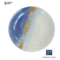 The Marie Daâge Horizon Gold Dinnerware features a round porcelain plate with a hand-painted blue and white gradient, accented by a gold streak. "Marie Daâge" logo in the top left; "27 Horizon Gold" in the bottom right.