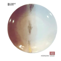 The Marie Daâge Horizon Gold Dinnerware features a round porcelain plate with a vertical gold, brown, and white gradient design and hand-painted details. The Marie Daâge logo appears in the top left. Made in France.