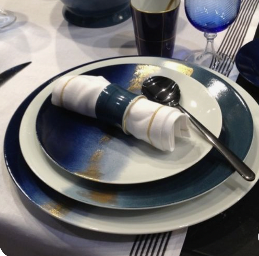 A table setting with Marie Daâge Horizon Gold Dinnerware, featuring blue and white gradient plates, a white napkin in a blue ring, and a spoon on top. Glassware and other French tableware appear in the background on a white tablecloth.