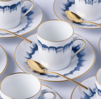 Marie Daâge Iris 2 Dinnerware features hand-painted white porcelain teacups and saucers with blue watercolor patterns, gold trim, and a matching gold spoon—capturing the luxury and elegance of Marie Daâge tableware.