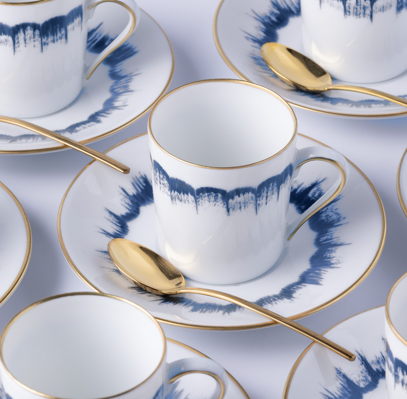 Marie Daâge Iris 2 Dinnerware features hand-painted white porcelain teacups and saucers with blue watercolor patterns, gold trim, and a matching gold spoon—capturing the luxury and elegance of Marie Daâge tableware.