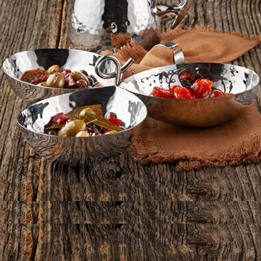 Three shiny metal bowls from the Mary Jurek Galaxy 3 Bowl Server, filled with assorted olives, pickled vegetables, and red peppers, rest on a rustic wooden table beside a brown cloth and a hammered stainless steel teapot.