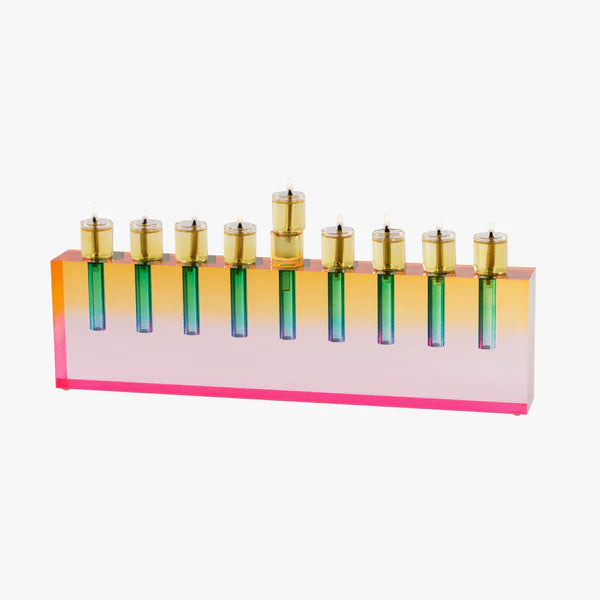 The Apeloig Modern Acrylic Oil Menorah features a rectangular base in gradient pink, orange, and yellow with nine gold candle holders arranged in a row, all set against a white background.