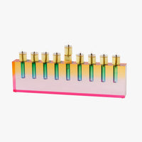 The Apeloig Modern Acrylic Oil Menorah features a rectangular base in gradient pink, orange, and yellow with nine gold candle holders arranged in a row, all set against a white background.