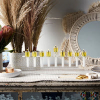 The Apeloig Modern Acrylic Oil Menorah with seven burning candles sits on a textured runner, accented by dried reeds, a macrame mirror in the background, and a flower vase for extra charm.