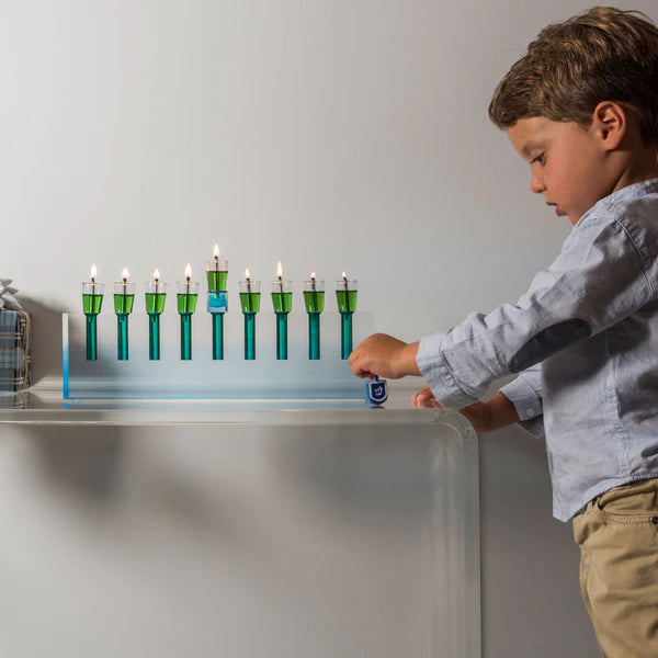 A young boy spins a dreidel beside the Apeloig Modern Acrylic Oil Menorah, glowing with nine lit candles on a clear table near a flower vase, celebrating Hanukkah.