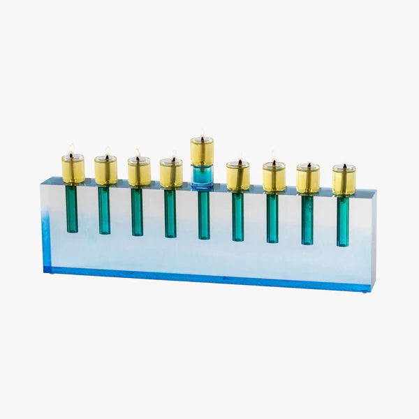 The Apeloig Modern Acrylic Oil Menorah features a blue rectangular base with nine gold and blue candle holders, each holding a lit candle. This customizable piece boasts a sleek, contemporary design perfect for any celebration.