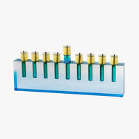 The Apeloig Modern Acrylic Oil Menorah features a blue rectangular base with nine gold and blue candle holders, each holding a lit candle. This customizable piece boasts a sleek, contemporary design perfect for any celebration.