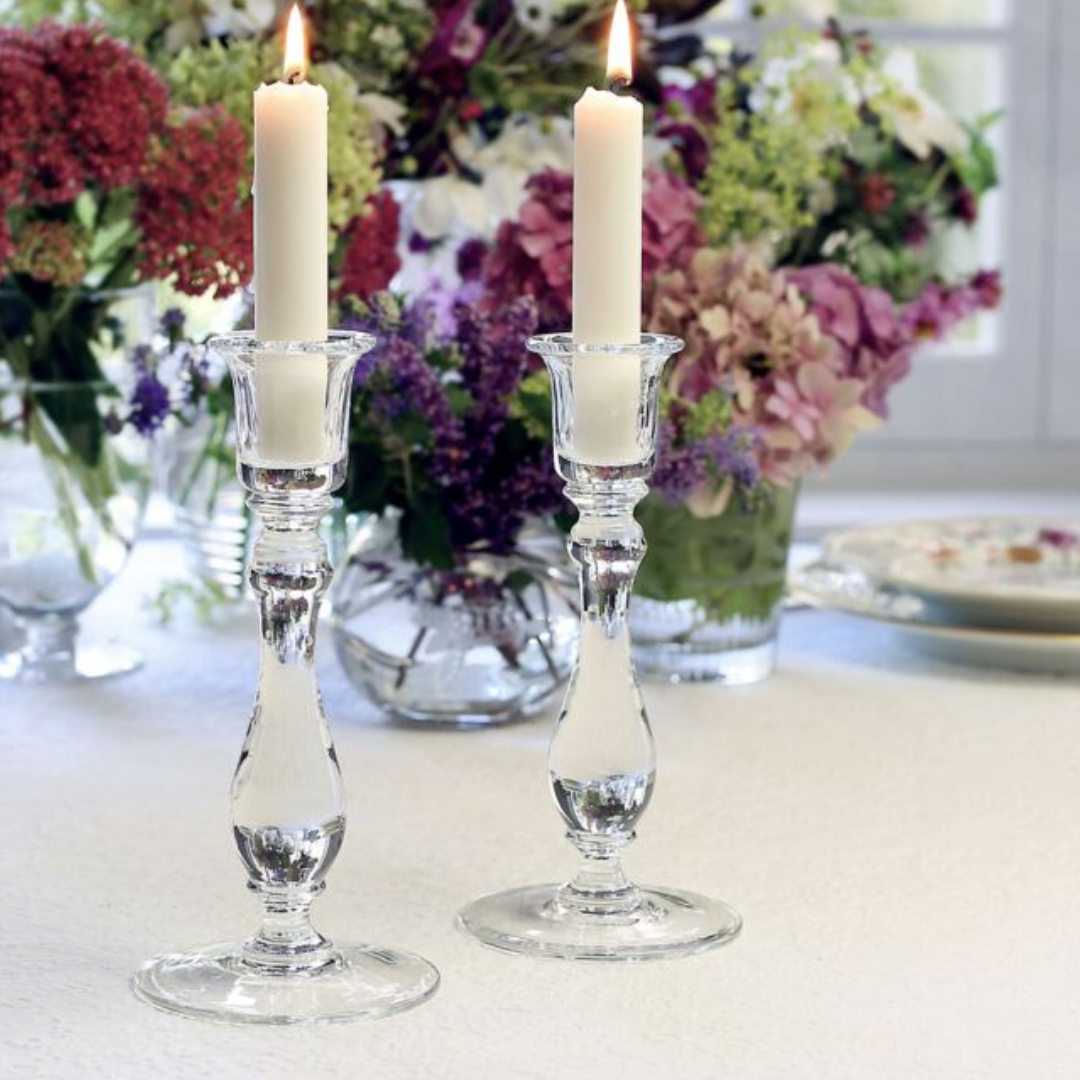 The Meryl Candlesticks Set of 2 by William Yeoward, each holding a lit white candle, sit on a white tablecloth amid floral arrangements and a window, creating an elegant, serene table setting ideal for refined home décor.