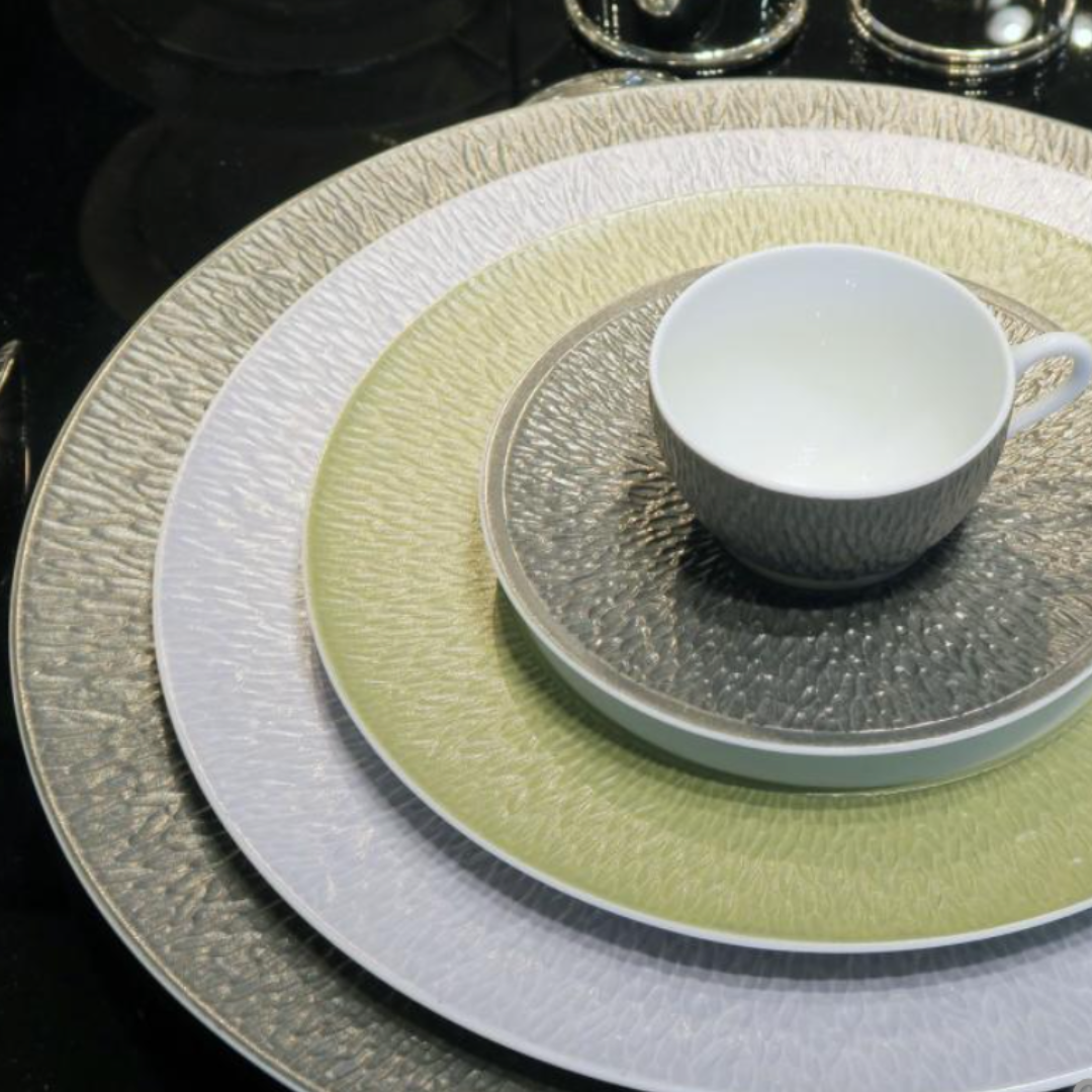 A white teacup on a matching saucer rests atop three stacked Raynaud Mineral Irise Dinnerware plates in Celadon—white, light green, and gray Limoges porcelain—arranged neatly on a reflective black surface.