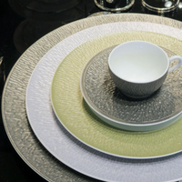 A white teacup on a matching saucer rests atop three stacked Raynaud Mineral Irise Dinnerware plates in Celadon—white, light green, and gray Limoges porcelain—arranged neatly on a reflective black surface.