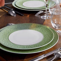 A close-up of a dining table set with Raynaud Mineral Irise Dinnerware - Celadon, featuring green-rimmed plates, silver cutlery, and crystal glassware on a geometric wooden table for an elegant meal setting.