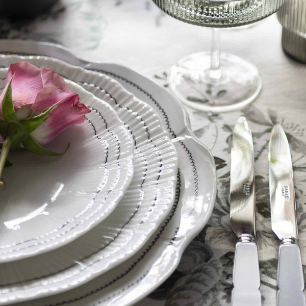 A close-up of a table set with Molecot's Coral Dinnerware Black plates stacked, a pink rose on top, silver cutlery, and a textured glass, all arranged on a floral tablecloth.