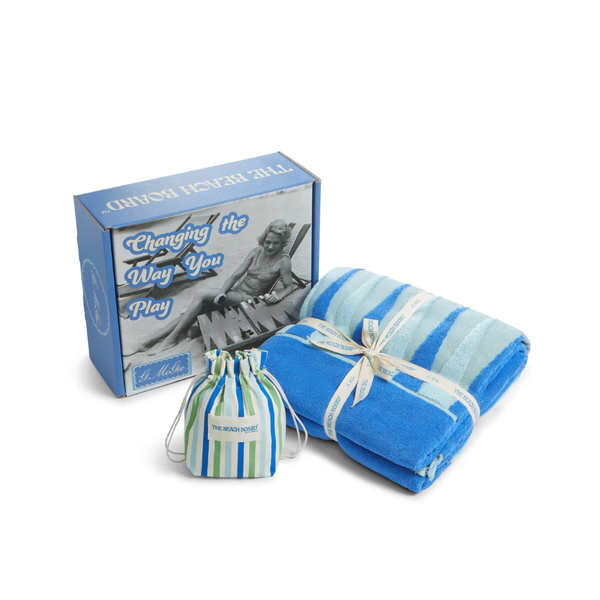 A blue G. McGee Beach Montauk Backgammon box with a vintage beach photo and "Changing the Way You Play" text sits next to a folded luxury terrycloth towel tied with ribbon and a small striped drawstring bag.