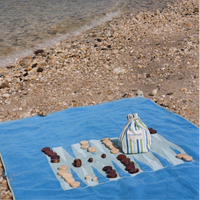 The G. McGee Beach Montauk Backgammon—a luxe terrycloth towel featuring a backgammon board, playing pieces, and striped bag—rests on pebbles by the clear shore.