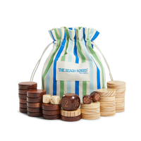 A striped blue, green, and white drawstring bag labeled “Beach Montauk Backgammon” by G. McGee sits behind stacks of light and dark wooden game pieces and dice, all arranged on a soft organic cotton beach towel.
