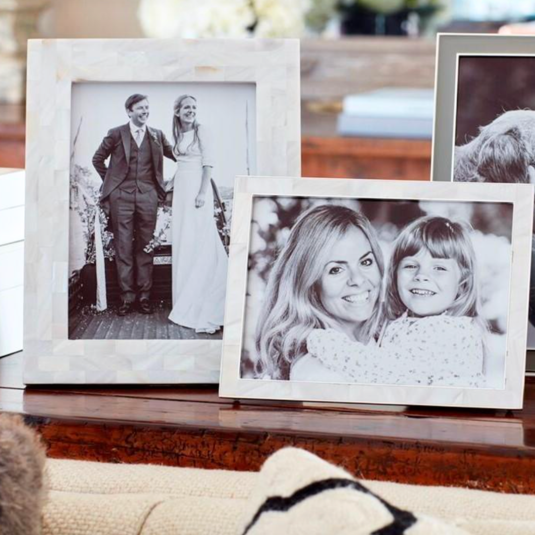 Two framed black-and-white photos sit on a wooden table: one in an Addison Ross Mother Of Pearl White Frame shows a couple in wedding attire, and the other displays a woman hugging a smiling young girl.