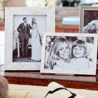 Two framed black-and-white photos sit on a wooden table: one in an Addison Ross Mother Of Pearl White Frame shows a couple in wedding attire, and the other displays a woman hugging a smiling young girl.