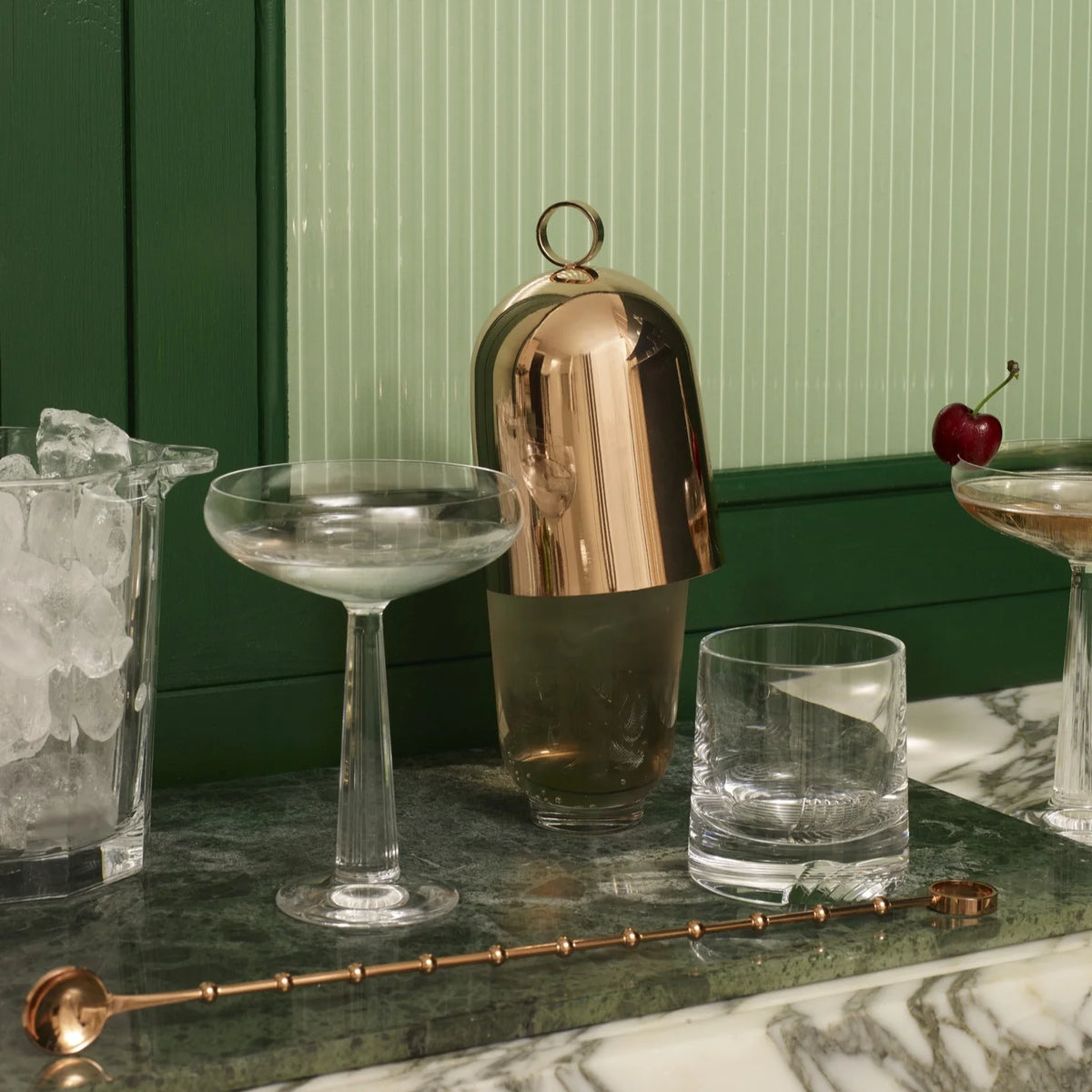 A marble bar counter displays a NUDE Glassware Big Top Coupe Glass from the Big Top Coupe Glasses Set of 2, a rocks glass, gold-capped cocktail shaker, glass of ice, cherry-garnished stirred drink, and copper bar spoon before a green paneled wall.