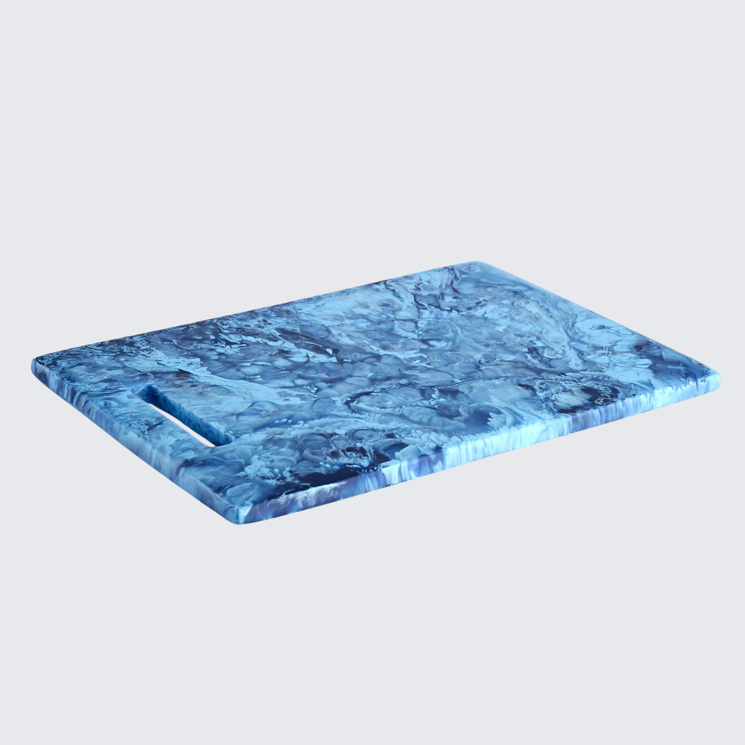 The Nashi Home Swirl Colored Resin Rectangle Chopping Board, featuring blue marble patterns and a small handle slot in the top left corner, is displayed on a light gray background.