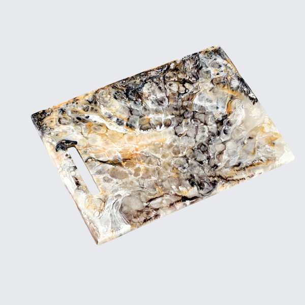 The Nashi Home Swirl Colored Resin Rectangle Chopping Board features a marbled design in black, white, beige, and brown with a left-side cutout handle, displayed on a light gray background.