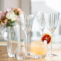 Vietri's Natalia High Ball glasses are arranged on a wooden table, some filled with drinks. One holds a yellow beverage with an orange slice garnish. An out-of-focus vase of flowers appears in the background.