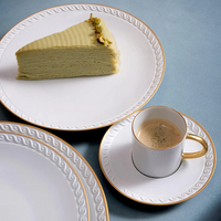 A slice of layered green cake with nuts rests on a white Neptune Gold plate by L’Objet, trimmed in 24k gold, beside a cup of coffee in a matching fine porcelain saucer, all atop a light blue surface.