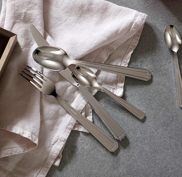 The Degrenne Normandy Flatware 5 Piece Set is arranged on a light pink cloth napkin on a gray countertop, with a Maison Degrenne wooden box partially visible in the top left corner.