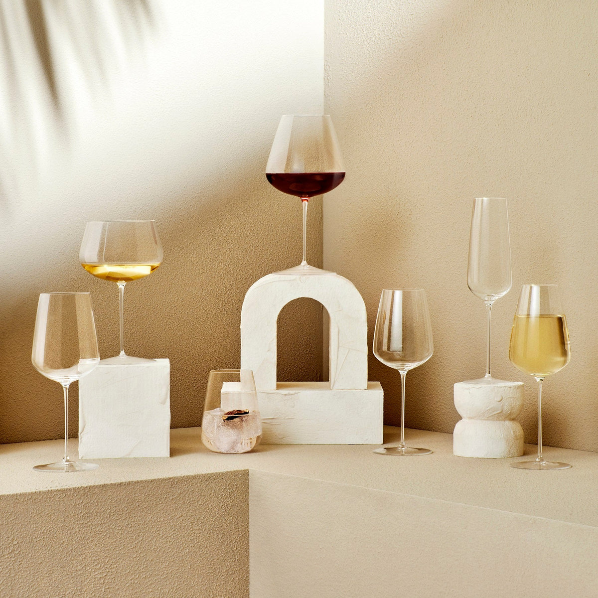 A pair of Nude Glassware’s Stem Zero Vertigo Water Glasses, crafted from lead-free crystal, is displayed on geometric white stone pedestals against a beige background with soft shadows.