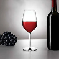 A glass of red wine, a dark wine bottle, and black grapes rest on a table against a gray backdrop—perfect for highlighting your Nude Glassware Climats All Purpose Wine & Water Glass Set of 2.