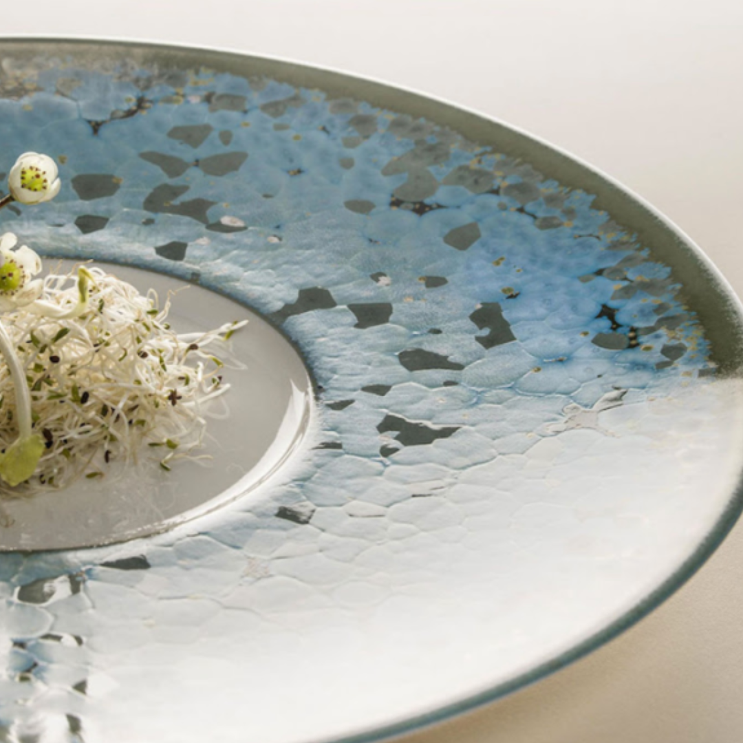 A close-up of a round Nymphea Dinnerware plate by Jaune De Chrome, made of Limoges porcelain with a blue textured glaze, featuring delicate white sprouts and green garnishes artfully arranged near the center.