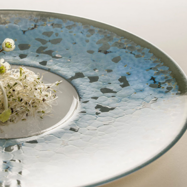 A close-up of a round Nymphea Dinnerware plate by Jaune De Chrome, made of Limoges porcelain with a blue textured glaze, featuring delicate white sprouts and green garnishes artfully arranged near the center.