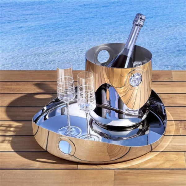 A bottle of champagne in an ice bucket with two empty flutes from the OH collection rests on the Oh De Stainless Steel Round Tray by Christofle, placed atop wooden boards with blue water in the background.