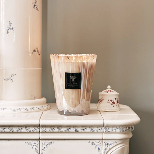 A Baobab Candle Pearls White sits on a white ceramic stove with blue floral designs, next to a small white jar with a red floral pattern, set against a light beige wall.