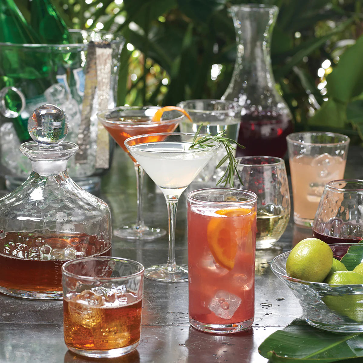 Colorful cocktails and spirits served in Juliska's Puro Glassware rest on a reflective outdoor table with greenery, limes, and dishwasher-safe glass decanters.