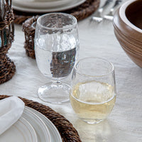Two Juliska Puro Glassware glasses—one with water, one with white wine—sit on a set table with woven placemats, plates, Portuguese linen napkins, and a wooden bowl. These textured glasses are also dishwasher safe.