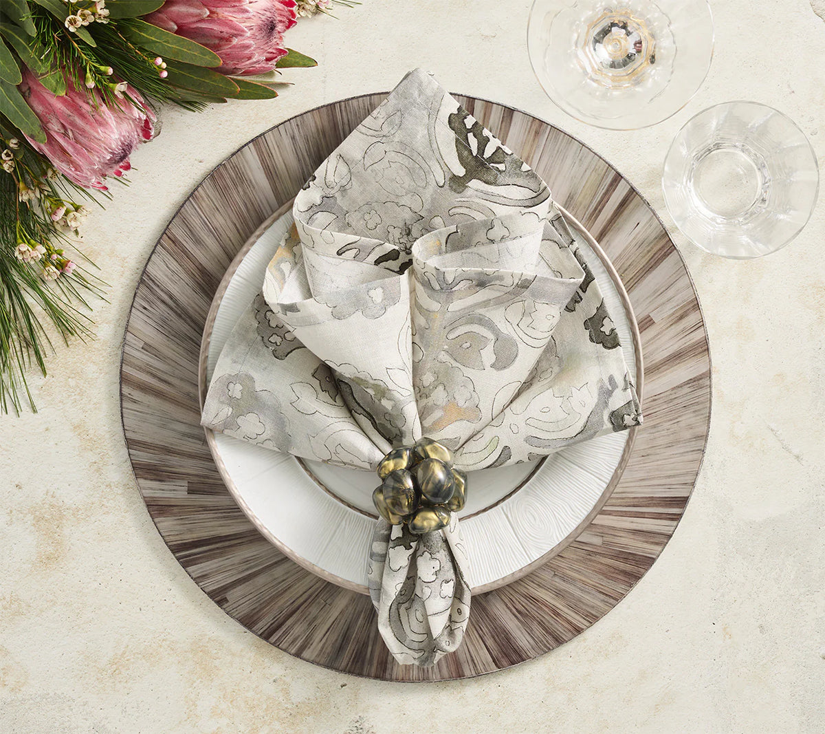 An elegant table is set with a patterned napkin in a ring on a white plate atop the Kim Seybert Aura Placemat Grey Set of 4, placed on a wooden charger, accented by wine glasses and a protea floral arrangement.