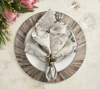 An elegant table is set with a patterned napkin in a ring on a white plate atop the Kim Seybert Aura Placemat Grey Set of 4, placed on a wooden charger, accented by wine glasses and a protea floral arrangement.