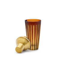 A tall, amber Prism Cocktail Shaker by L'Objet with vertical grooves stands upright next to a matching shaker lid, both displayed on a white background.