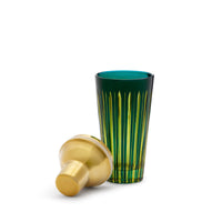 A tall, hand-crafted green glassware with vertical yellow stripes stands upright beside a gold Prism Cocktail Shaker lid by L’Objet, both displayed against a plain white background.
