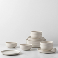 Costa Nova Pacifica Vanilla Dinnerware, featuring minimalist off-white ceramic plates and bowls, is stacked and arranged on a white surface against a plain light gray background.