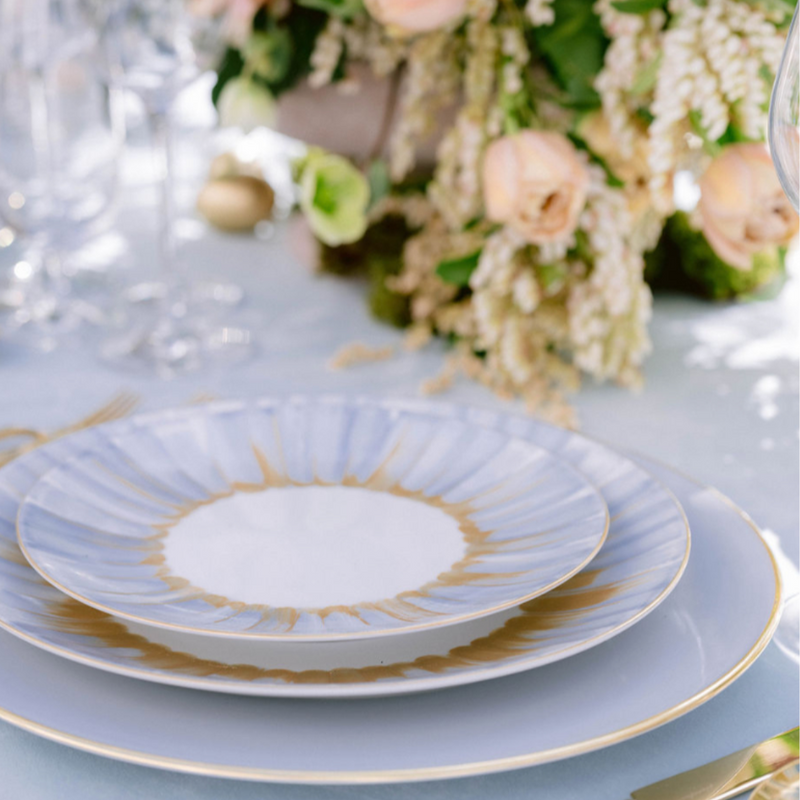 Marie Daâge Panache plates in white Limoges porcelain with gold and blue accents are arranged on a white tablecloth, complemented by pastel flowers and greenery in the background for an elegant table setting.