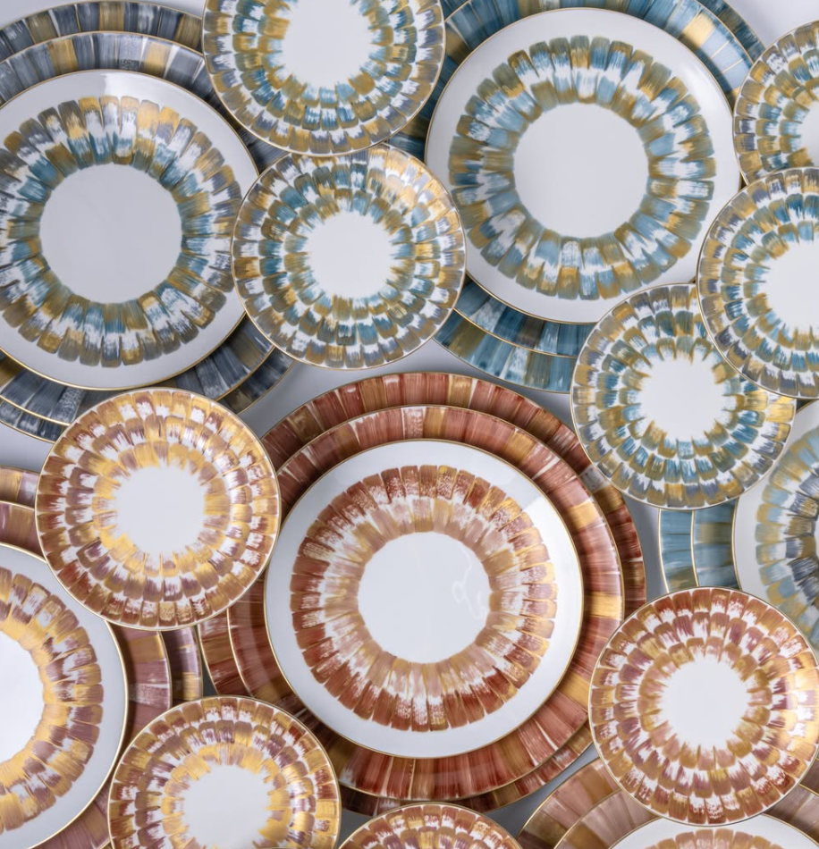 Marie Daage Panache plates by Marie Daâge feature circular, radiating patterns in blue, gold, and brown. Overlapped in a colorful arrangement, they offer a luxurious and symmetrical dinnerware display.