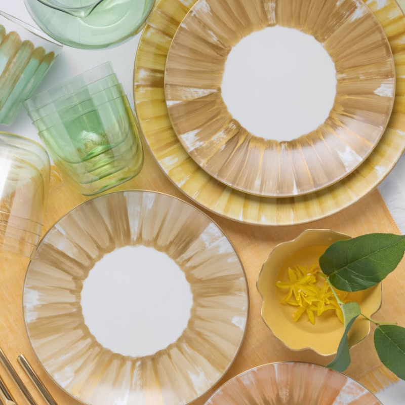 A flat lay of a table setting with Marie Daâge Panache luxury dinnerware, showcasing gold and white patterned plates, green glassware, and a small yellow dish adorned with yellow flowers and a green leaf.