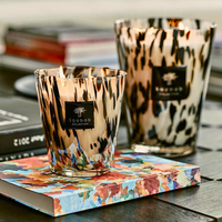 Two Baobab Pearls Candles, including the Black Pearls scent in hand-blown glass, sit atop a vibrant book with additional books and a blurred background on a dark tabletop—ideal for adding luxury home fragrance.