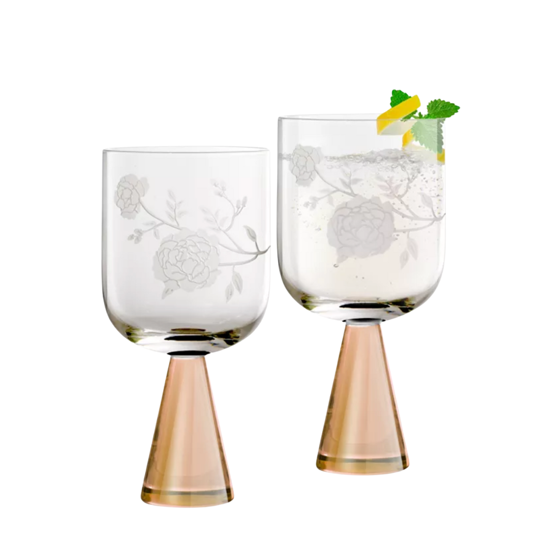 The Peony Goblet Set of 2 by Current Home features clear glassware with etched floral designs and rose gold stems. One handcrafted goblet is shown holding a drink garnished with lemon and mint.