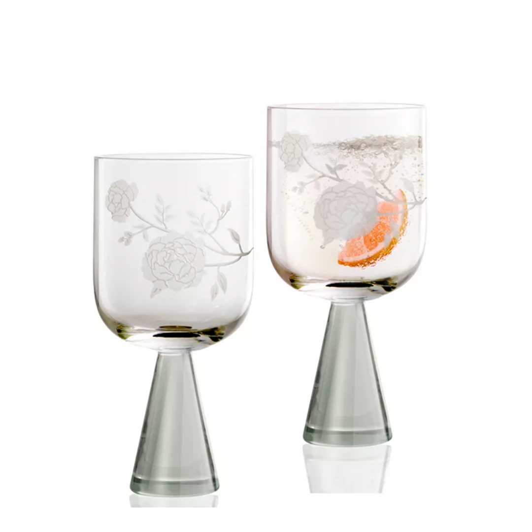 The Peony Goblet Set of 2 by Current Home features modern handcrafted glassware with etched floral designs and conical bases—one empty, the other with a drink, ice, and orange slice—set against a white background.