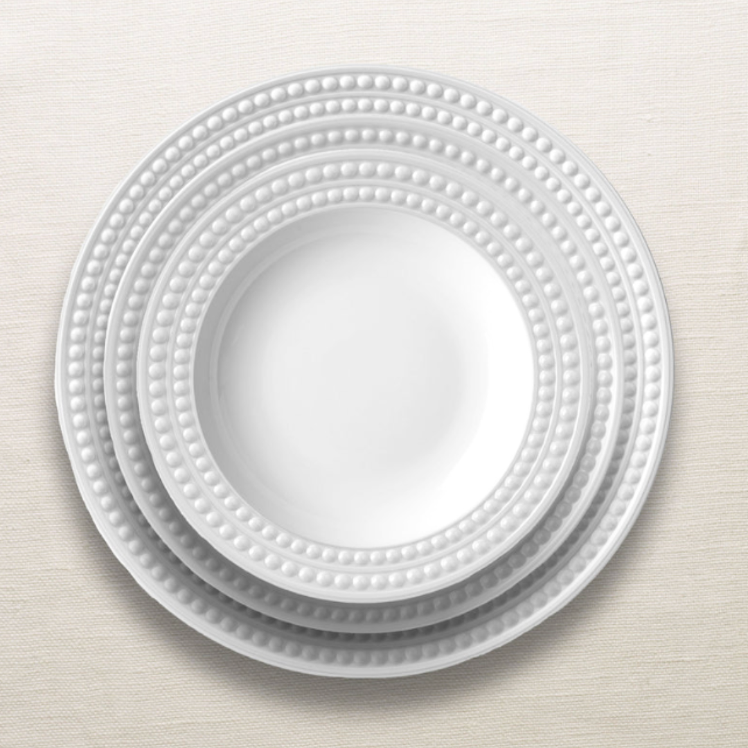 Four white ceramic plates from L'Objet's Perlèe White Dinnerware collection, each featuring a beaded rim, are neatly stacked on a light, textured surface.