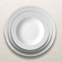 Four white ceramic plates from L'Objet's Perlèe White Dinnerware collection, each featuring a beaded rim, are neatly stacked on a light, textured surface.